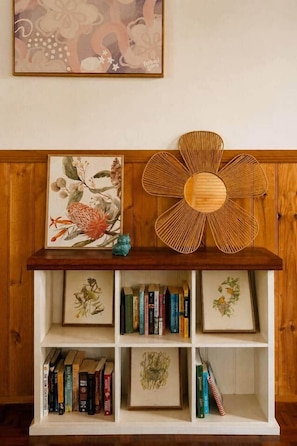 Ein Bücherregal mit gerahmten Pflanzenbildern und Büchern, einem gewebten Dekorationsobjekt und einem an der Wand hängenden abstrakten Gemälde.