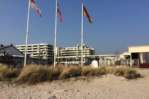 A building with multiple floors, a flagpole with flags, and a sandy foreground with grass. - Grömitz-Center II No. 31 (Grömitz)