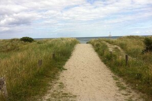 Ein sandiger Pfad führt zum Meer, gesäumt von Gras, unter einem bewölkten Himmel.