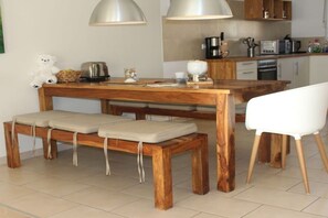Ein Holztisch mit Bänken, ein weißer Stuhl und Küchengeräte im Hintergrund.