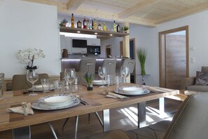 A dining area with a wooden table set for four, a kitchen in the background, and a door to the right. - Hochalmbahnen Chalet (Rauris)