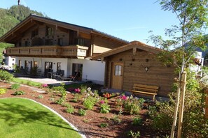 Un bâtiment à deux étages doté d’un balcon, d’un jardin avec différentes espèces de plantes et d’un banc.