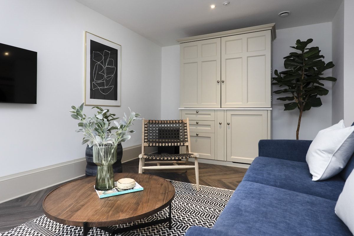 A living room with a blue sofa, a round coffee table, a rattan chair, a large plant, and a white cabinet.