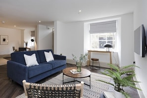 Uma sala de estar moderna com um sofá azul, uma mesa de centro redonda, um tapete estampado e uma planta de samambaia.