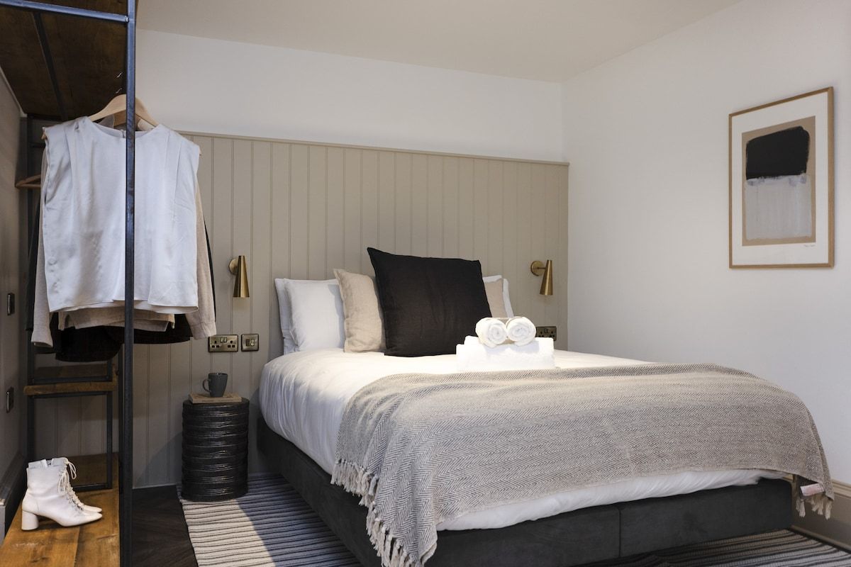 A bedroom with a bed, a hanging rack with clothes, a small round table with a cup, and a pair of boots.