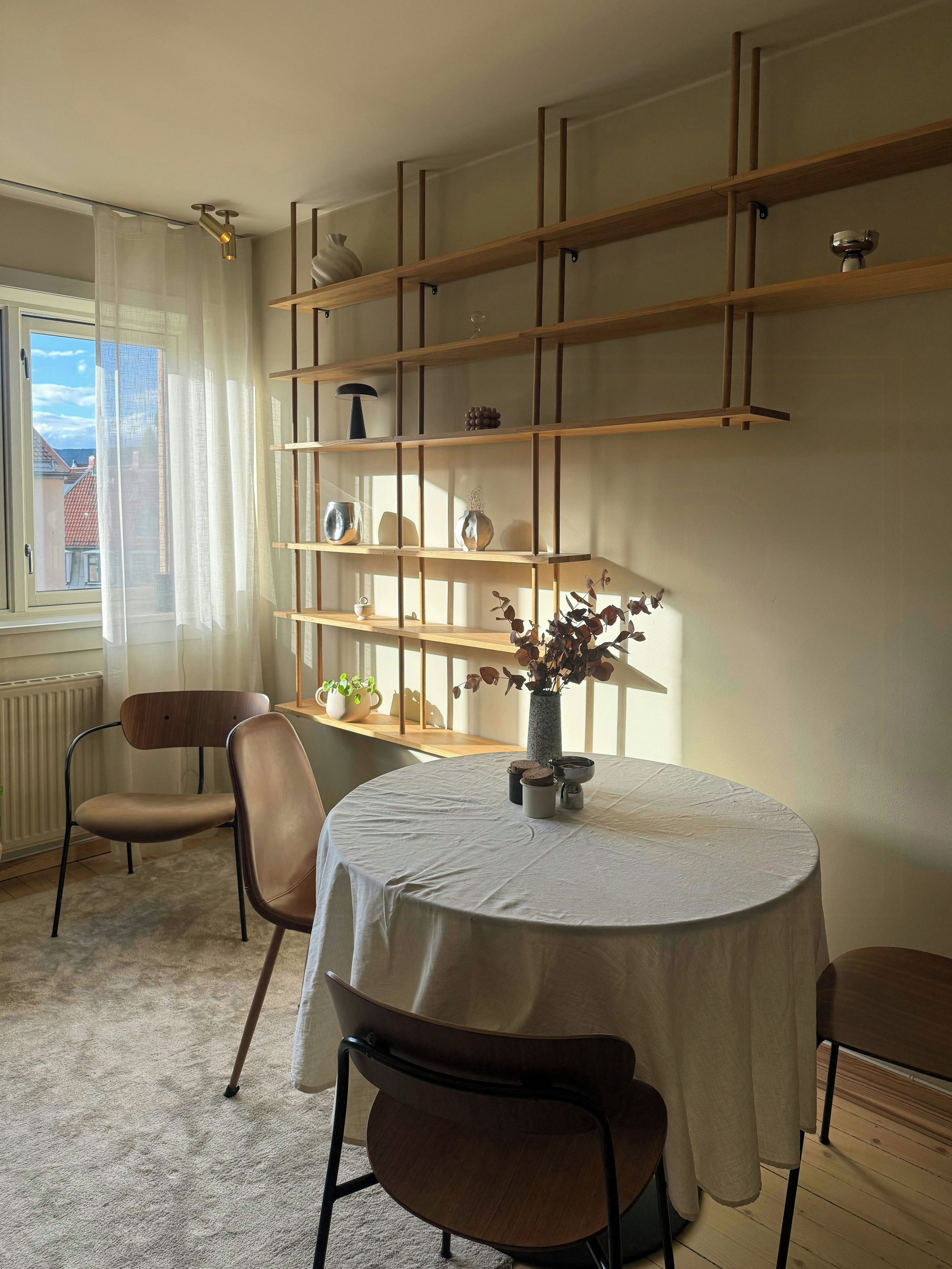 A room with a round table, two chairs, a bookshelf, and a window with curtains.