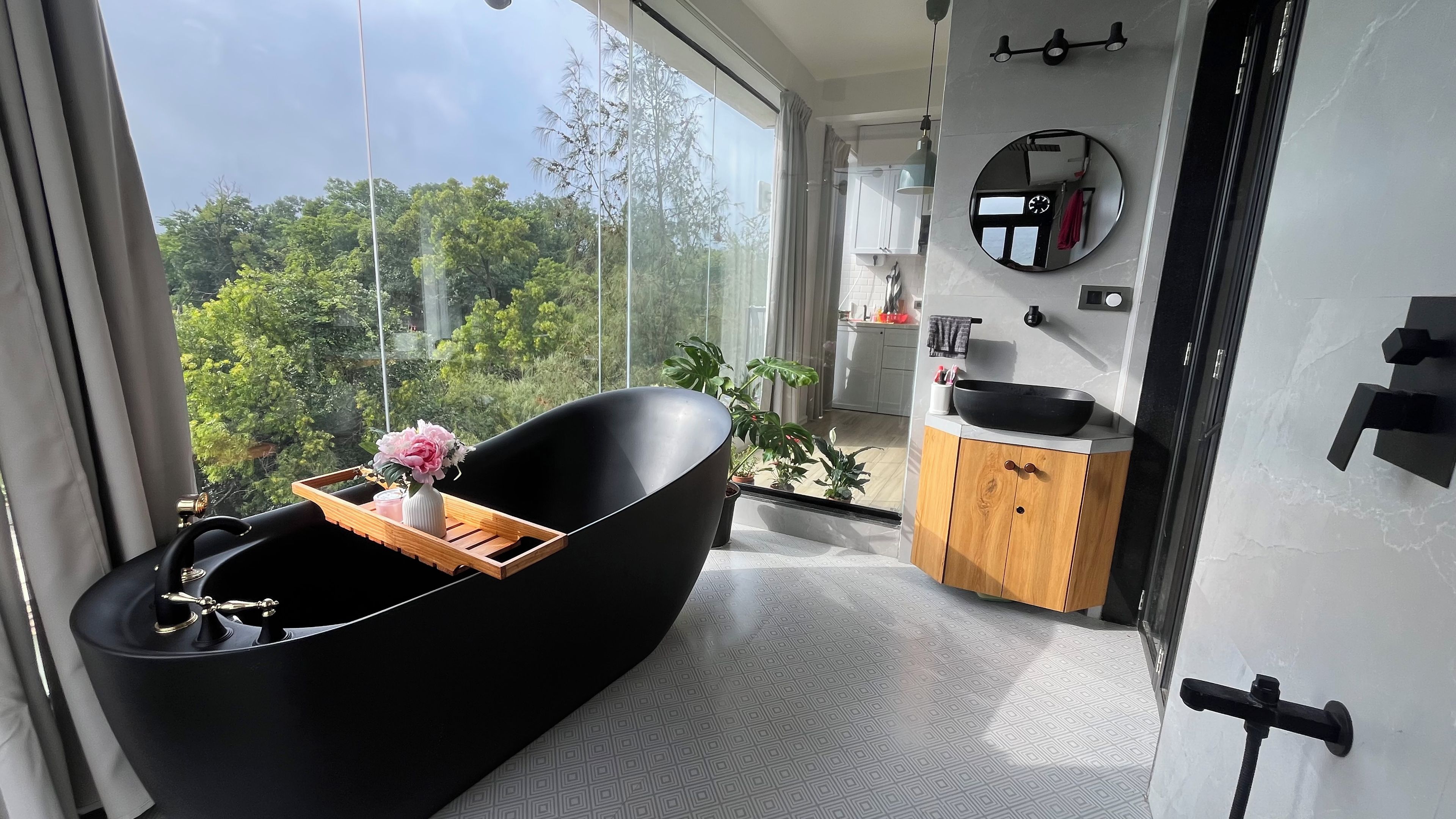 Een moderne badkamer met een zwart bad, een houten dienblad met een vaas en een kaars, en uitzicht op groen buiten.