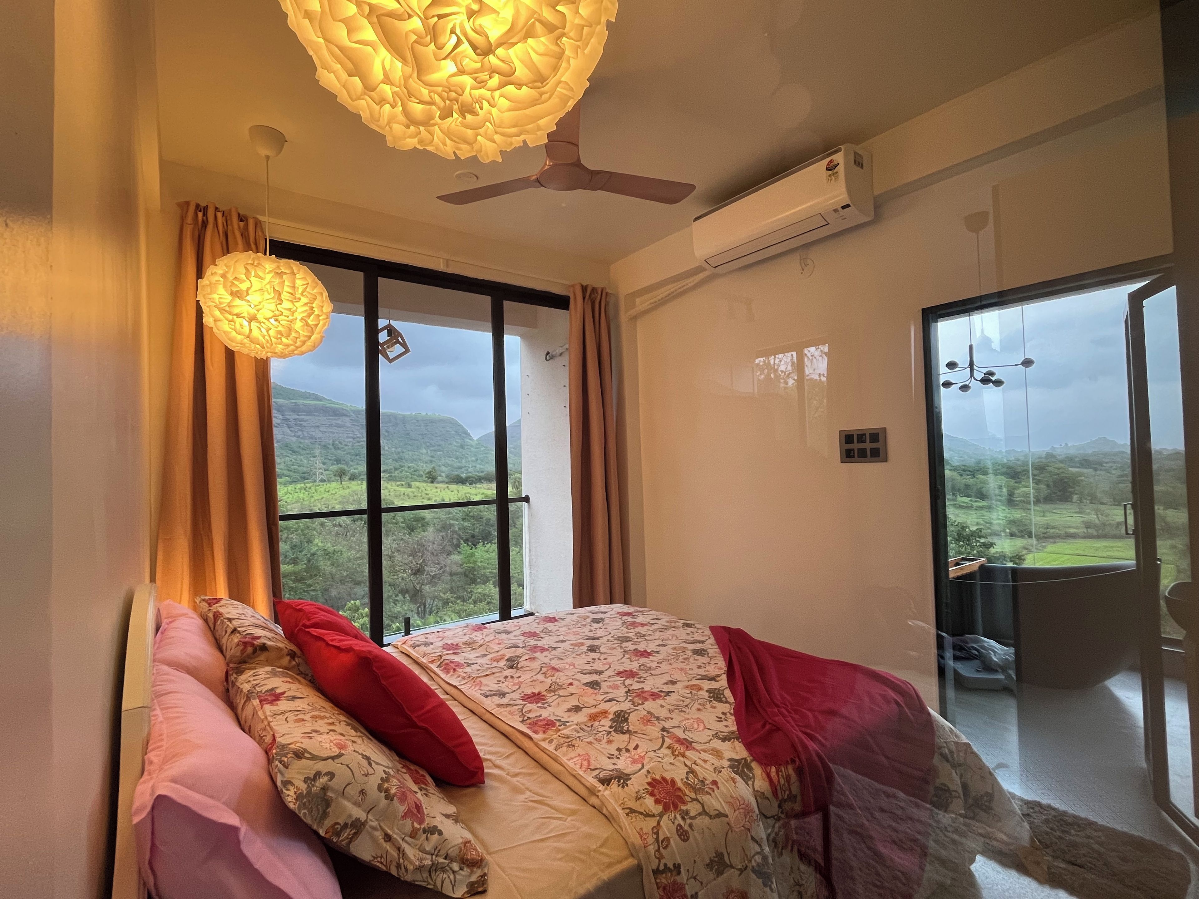 Een slaapkamer met een bed, gordijnen, een plafondventilator, een airconditioning en uitzicht op een landschap door een groot raam.