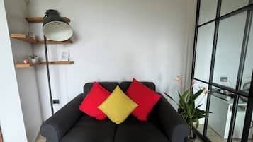 Una sala de estar moderna con un sofá oscuro, cojines rojos y amarillos, una lámpara de pie y una planta en maceta.