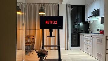 Une cuisine moderne avec un téléviseur à écran plat affichant le mot « NETFLIX » en lettres rouges, un coin repas avec un tabouret de bar et un vase rempli de fleurs posé sur le sol.