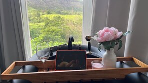 A wooden tray with a tablet, a glass, and a candle on a table by a window with a view of greenery.