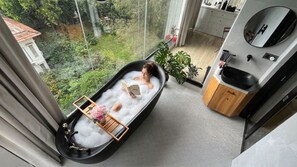 A room with a bathtub filled with bubbles, a wooden tray, and a book. There is a view of greenery outside the window.