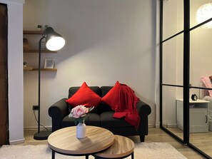 A modern living room with a black sofa, red cushions, a white vase with flowers, a round wooden coffee table, and a floor lamp.