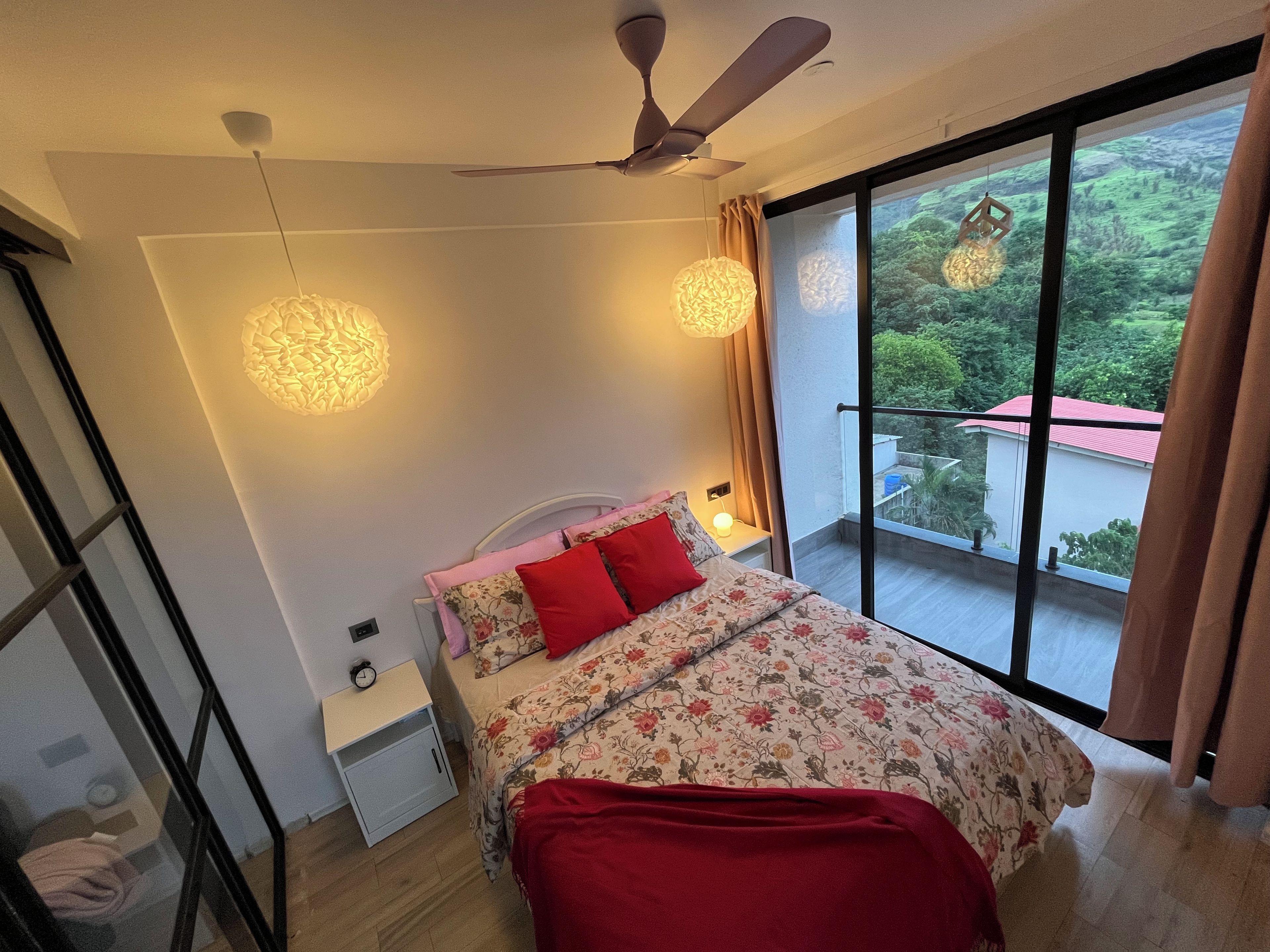 Quarto com cama, mesa de cabeceira, abajur, ventoinha no teto e vista para o verde através de uma janela grande.