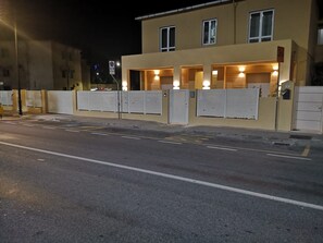 Um edifício com entrada protegida por grades, área de estacionamento e uma rua em primeiro plano.