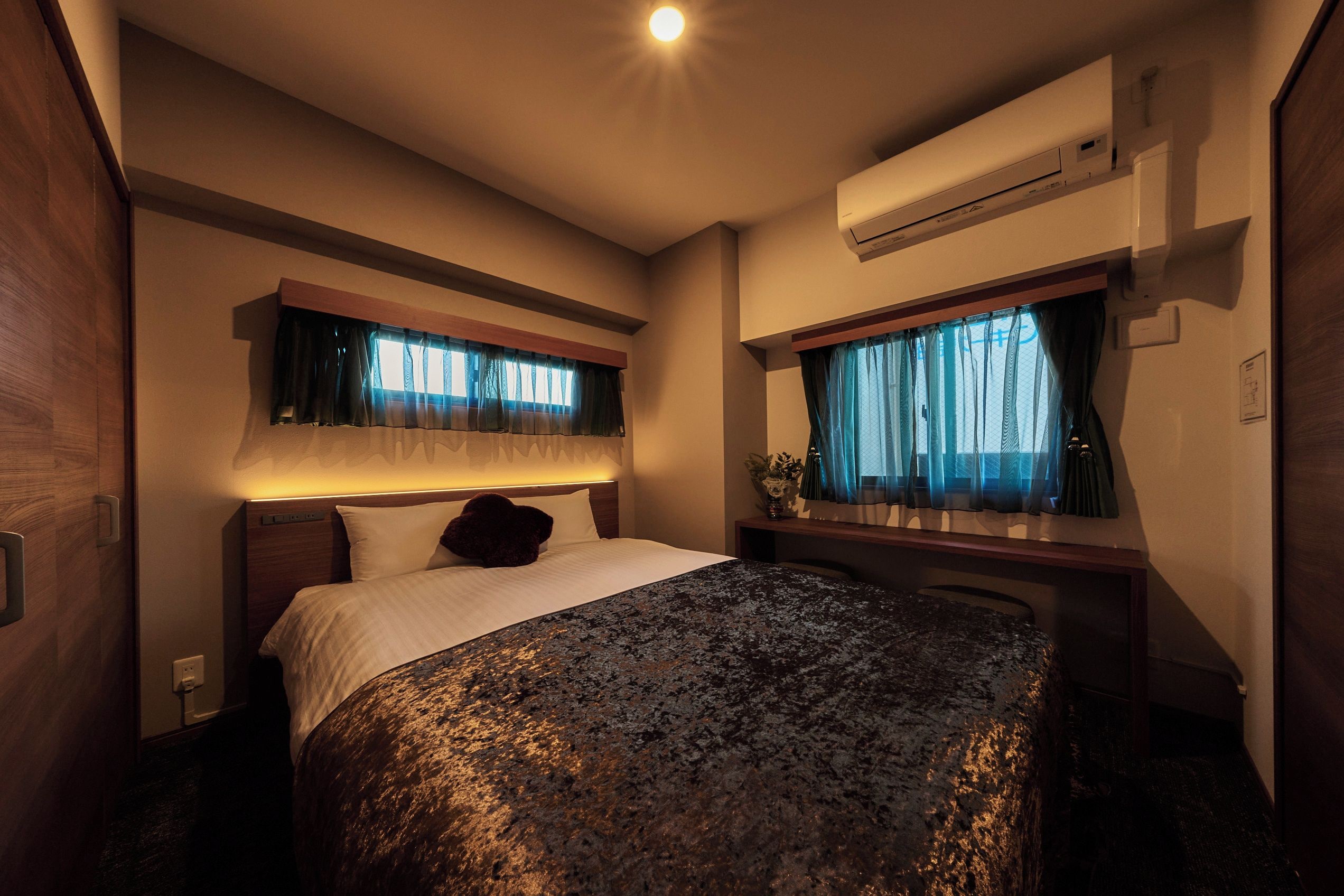 Habitación de hotel con cama, equipo de aire acondicionado, ventana con cortinas y una pequeña mesa con un florero.