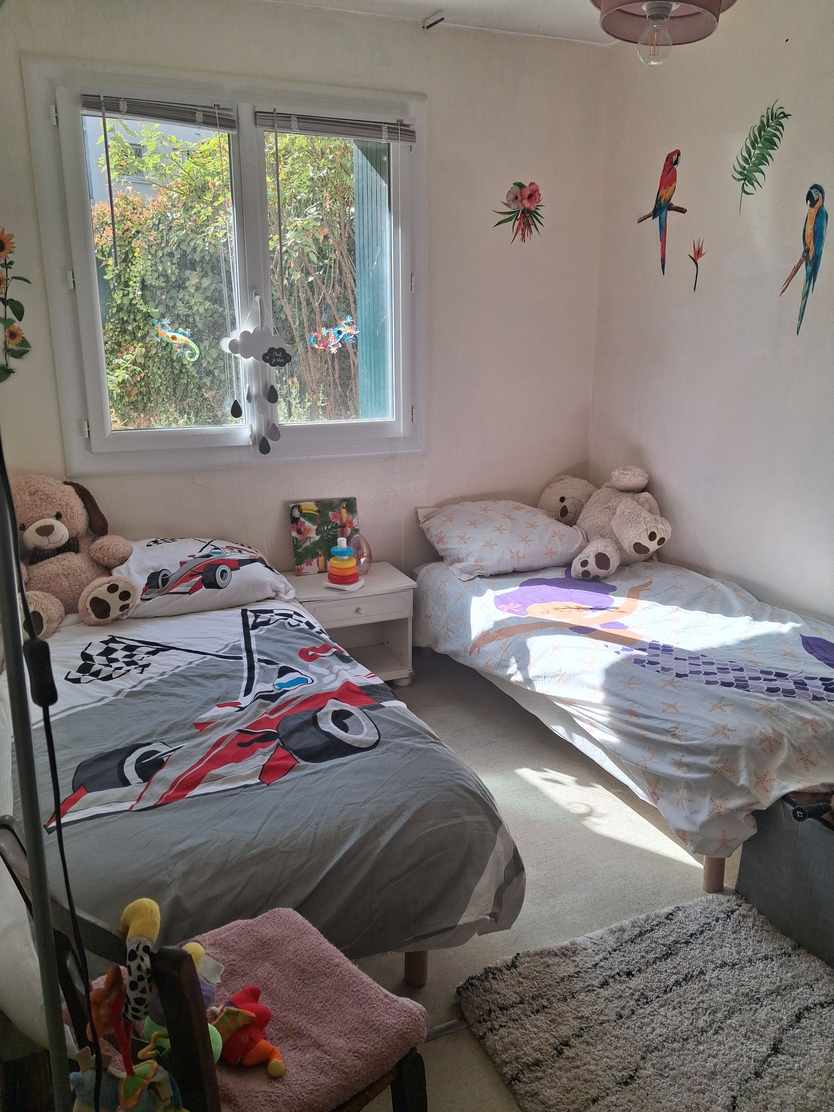 Een slaapkamer met twee bedden, een raam met uitzicht op groen, een muur met een papegaai en plantdecoratie, en een teddybeer aan de linkerkant.