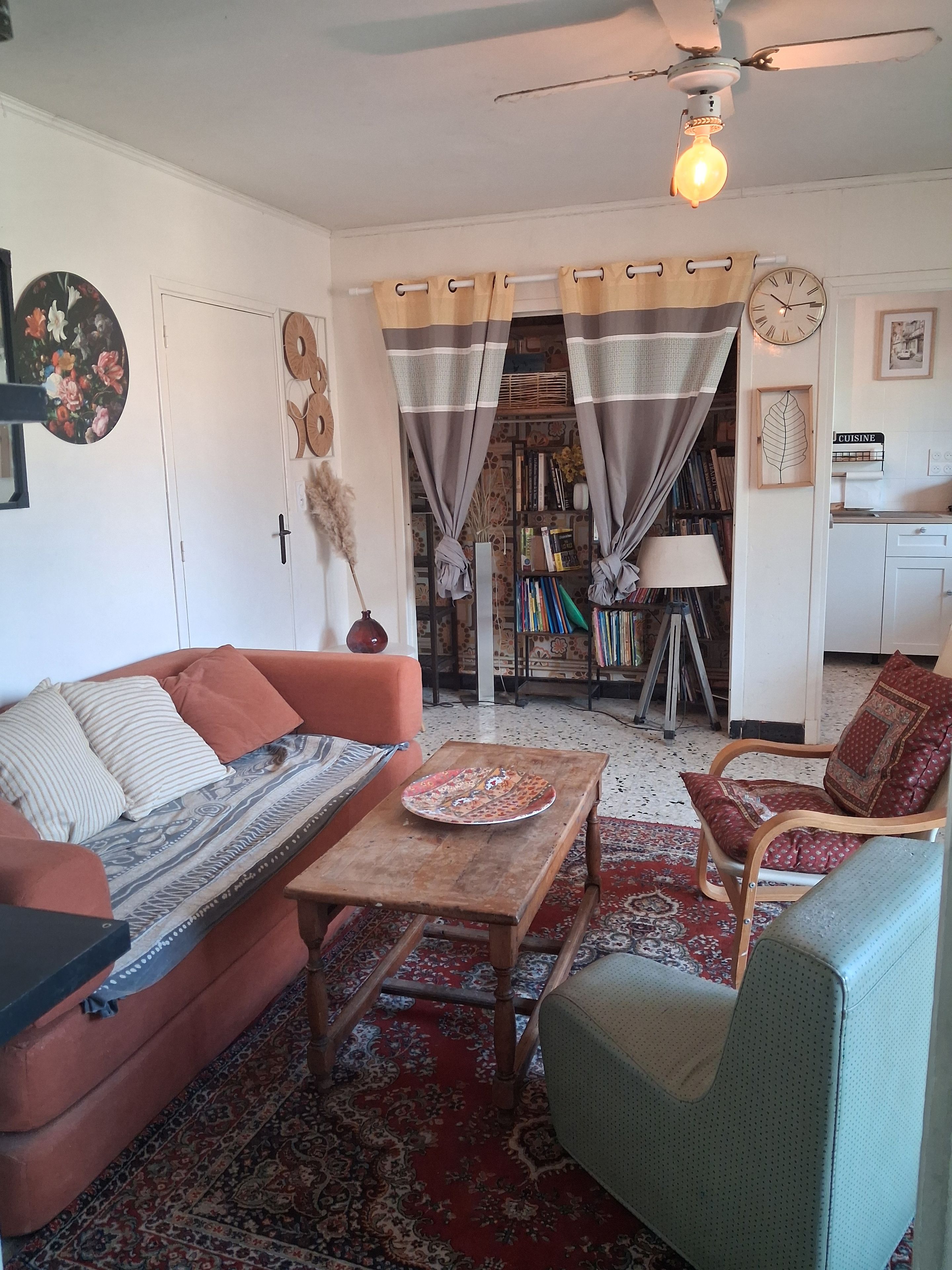 Een gezellige woonkamer met een bank, fauteuil en een salontafel. Er is een boekenplank, een klok en een plafondventilator.
