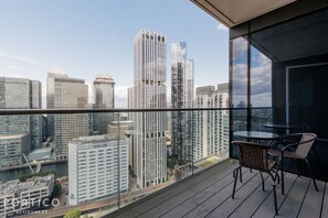 Un balcón con vista a un paisaje urbano, que incluye edificios altos y un río. Hay una mesa y dos sillas en el balcón.