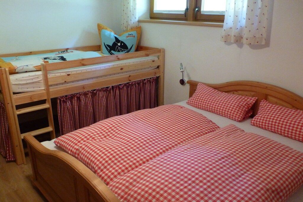 A bedroom with a bunk bed, a single bed, checkered bedding, a window with curtains, and a wall-mounted light.