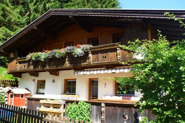 Un chalet traditionnel en bois, doté d’un balcon orné de fleurs, d’une clôture en bois et offrant une vue sur une zone boisée.