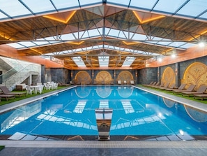 A swimming pool with a geometric ceiling structure, surrounded by lounge chairs and a stone wall with decorative panels. - Qorqud Hotel - Free Breakfast & Dinner Included (Baku)