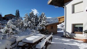Un espacio al aire libre cubierto de nieve, con un banco de madera y una mesa de picnic, un edificio con una ventana y árboles cargados de nieve.