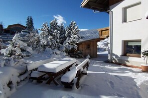 Un espace extérieur enneigé avec un banc en bois et une table de pique-nique, un bâtiment doté d’une fenêtre et des arbres couverts de neige.