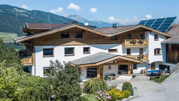 Un chalet de dos plantas con techo inclinado, paneles solares y un balcón con mobiliario de exterior.