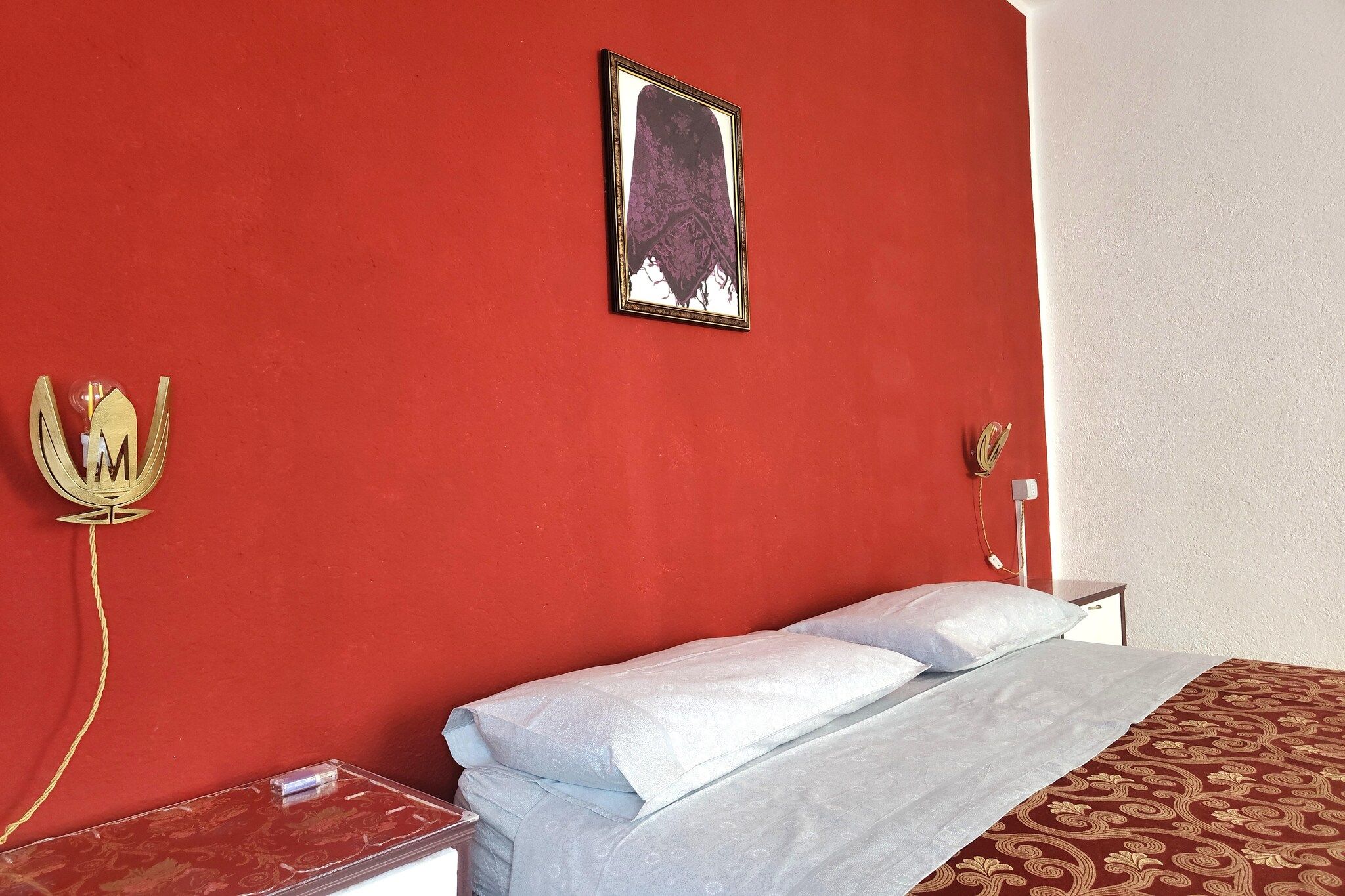 Ein Hotelzimmer mit roter Wand, einem Bett mit weißer Bettwäsche, einem gerahmten Bild, einem dekorativen Gegenstand, der einer Krone ähnelt, und einem Nachttisch mit einer Lampe.