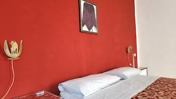Ein Hotelzimmer mit roter Wand, einem Bett mit weißer Bettwäsche, einem gerahmten Bild, einem dekorativen Gegenstand, der einer Krone ähnelt, und einem Nachttisch mit einer Lampe.