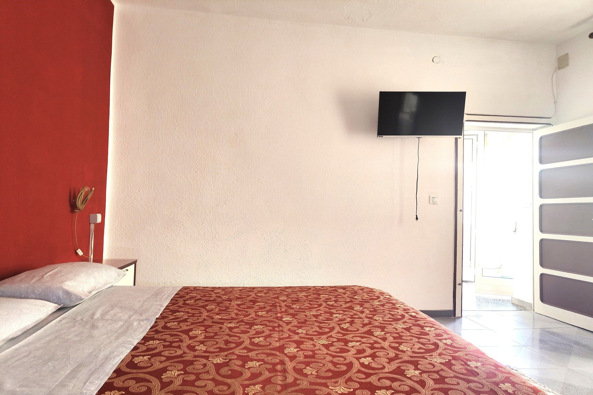 Una camera d'albergo con un letto, una televisione a parete e una porta che dà su un balcone.