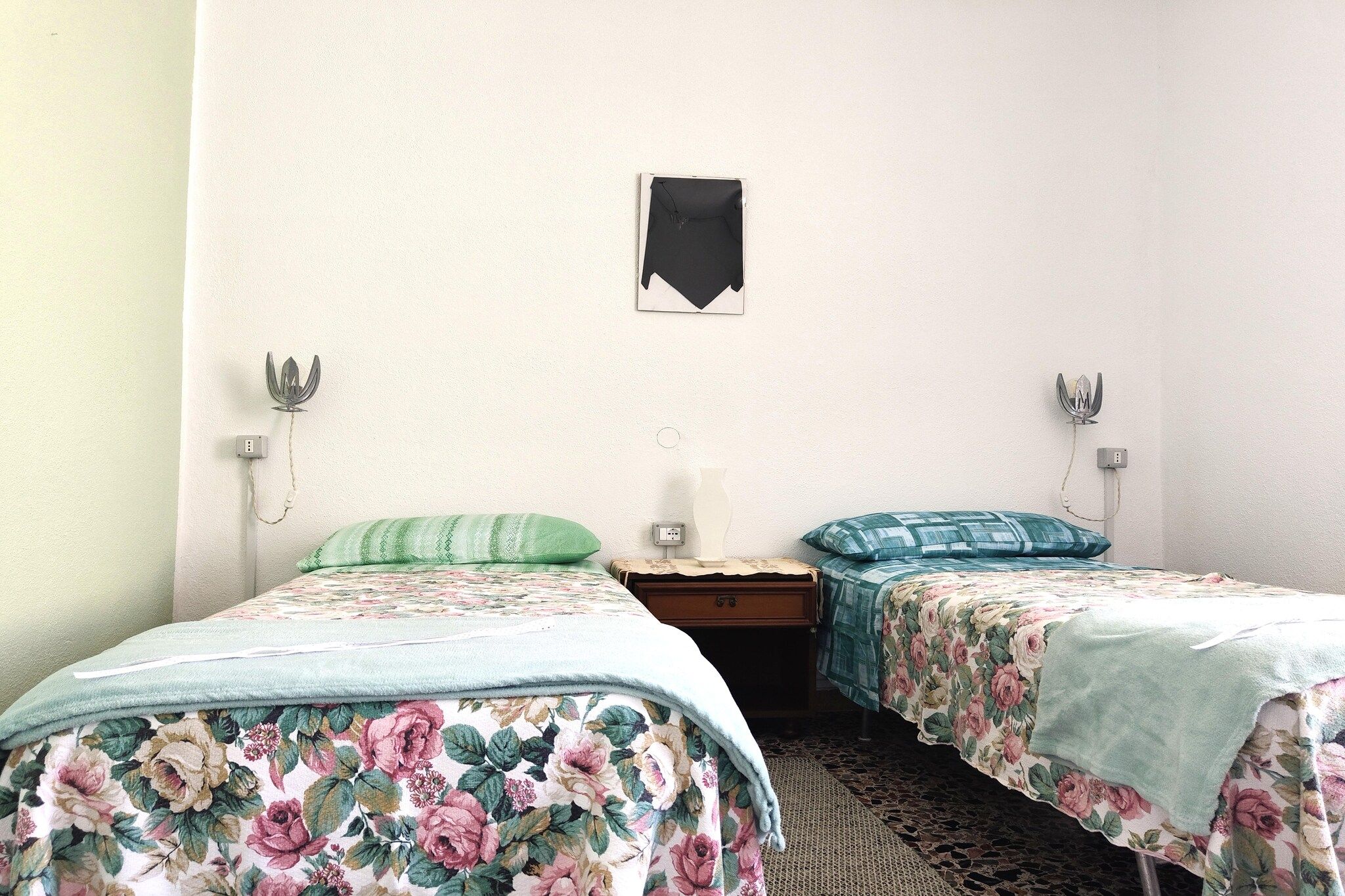 A room with two single beds, each with floral bedding, a small table, and wall-mounted lamps.