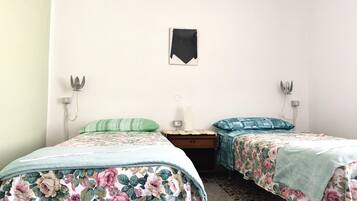 A room with two single beds, each with floral bedding, a small table, and wall-mounted lamps.