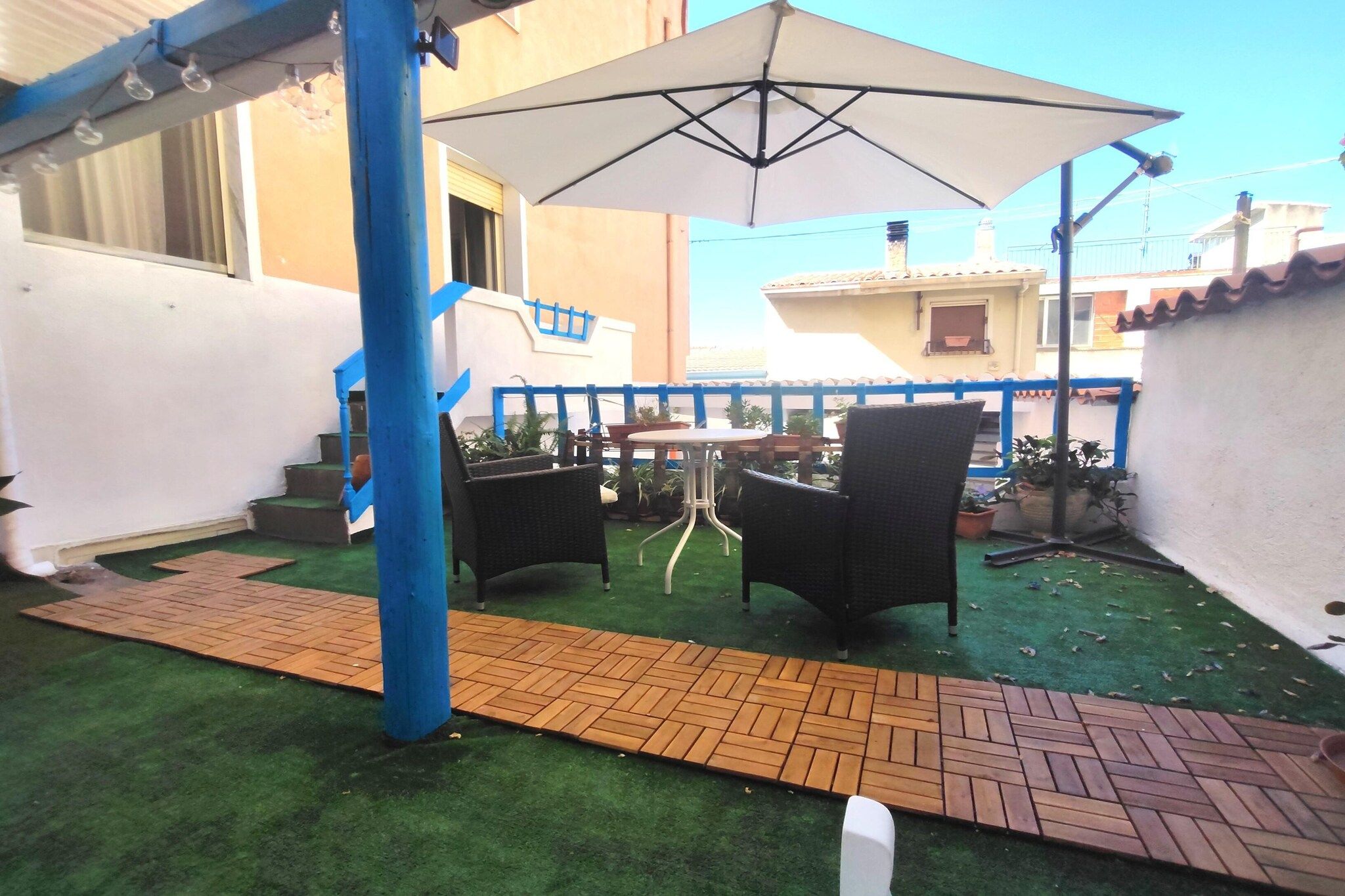 Patio esterno con erba sintetica, un vialetto in mattoni e due sedie in vimini sotto un ombrellone grande.