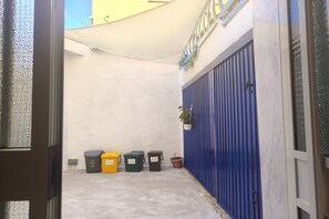 A narrow outdoor corridor with a blue door, three waste bins, and a potted plant.