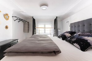 Quarto com cama, mesa de cabeceira, prateleira na parede e janela com cortinas.