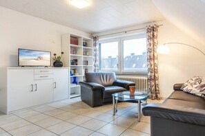 Ein modernes Wohnzimmer mit einer Couch, einem Couchtisch, einem Fernseher, einem Bücherregal und einem Fenster mit Vorhängen.