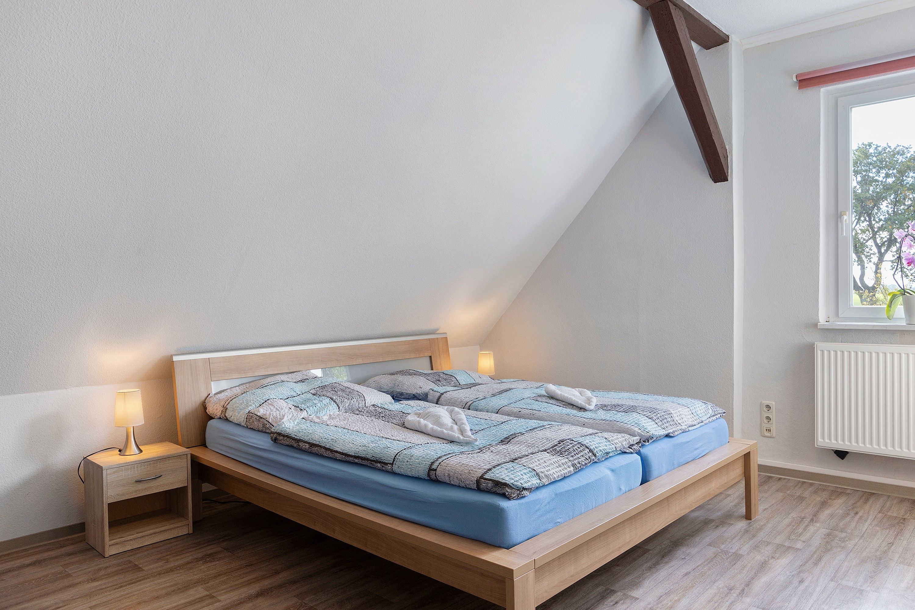 Ein Schlafzimmer mit schrägverdeckter Decke, einem hölzernen Bettgestell, einem Nachttisch mit Lampe und einem Fenster mit Blick auf Bäume.