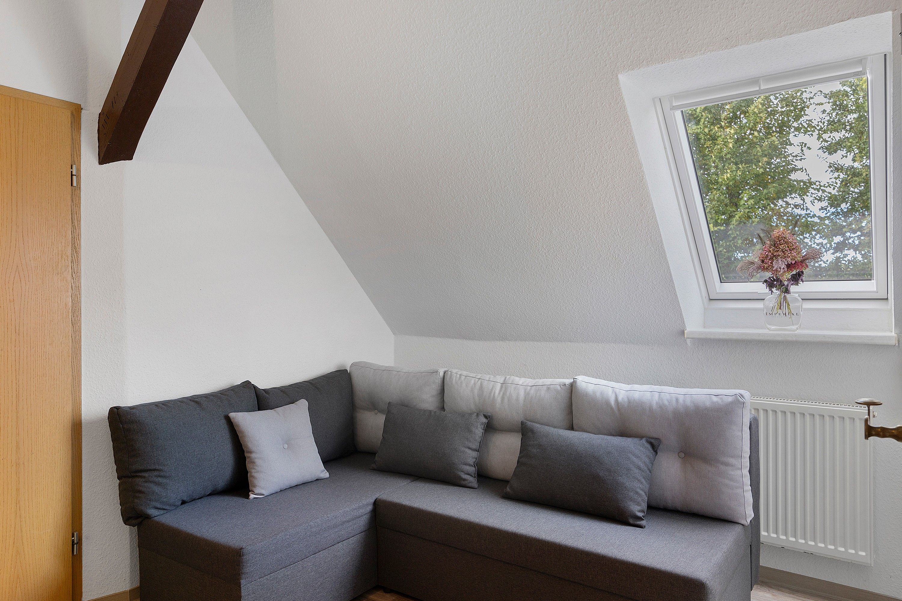 A corner room with a grey sofa, a window with a view of trees, and a white ceiling.