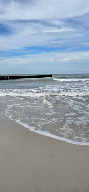 Une plage où les vagues viennent lécher le rivage, une jetée en bois qui s’avance dans l’eau et un ciel dégagé au-dessus.