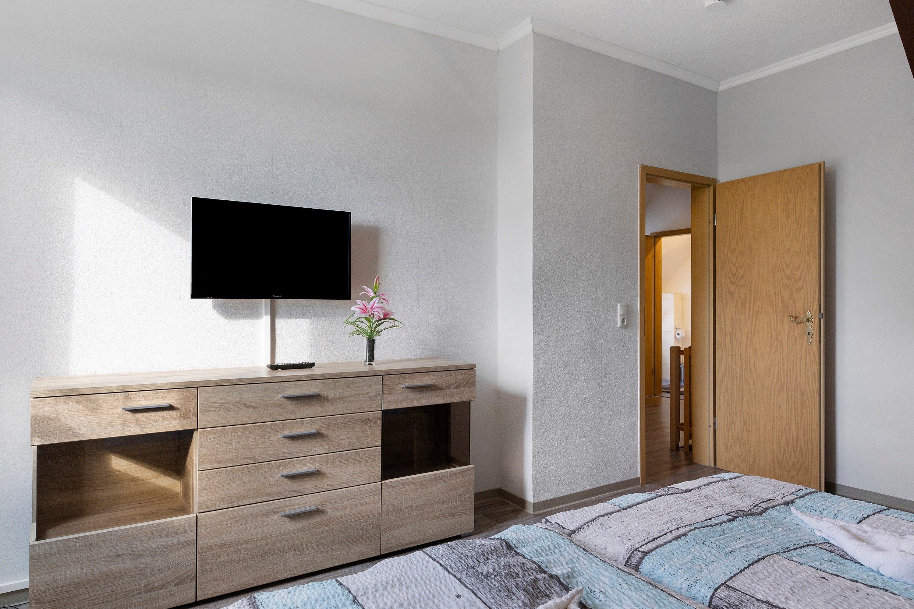Een slaapkamer met een bed, een televisie op een dressoir, een houten deur en een kleine vaas met bloemen.