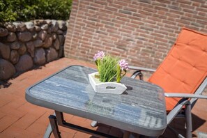 A glass-top table with a small planter containing pink flowers, positioned on a patio with a brick wall and a stone wall in the background. - 1 in the house "Trischen" (ID 359) (St. Peter-Ording)