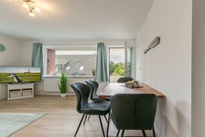 A dining area with a wooden table, black chairs, a potted plant, and a view of a brick building through a window. - Apartment 6 "Home Port" (ID 496) (St. Peter-Dorf)