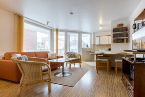 Ein modernes Wohnzimmer mit einer Couch, Sesseln, einem Esstisch und einer Küchenzeile mit Bücherregal.