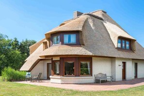 Ein Reetdachhaus mit Backsteinsockel, großen Fenstern und einer Terrasse mit Gartenmöbeln.