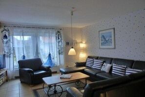 A living room with a sofa, armchair, and a coffee table. There is a window with curtains, a wall-mounted lamp, and a framed picture. - Holiday home in Graal-Müritz with terrace (Graal-Müritz)