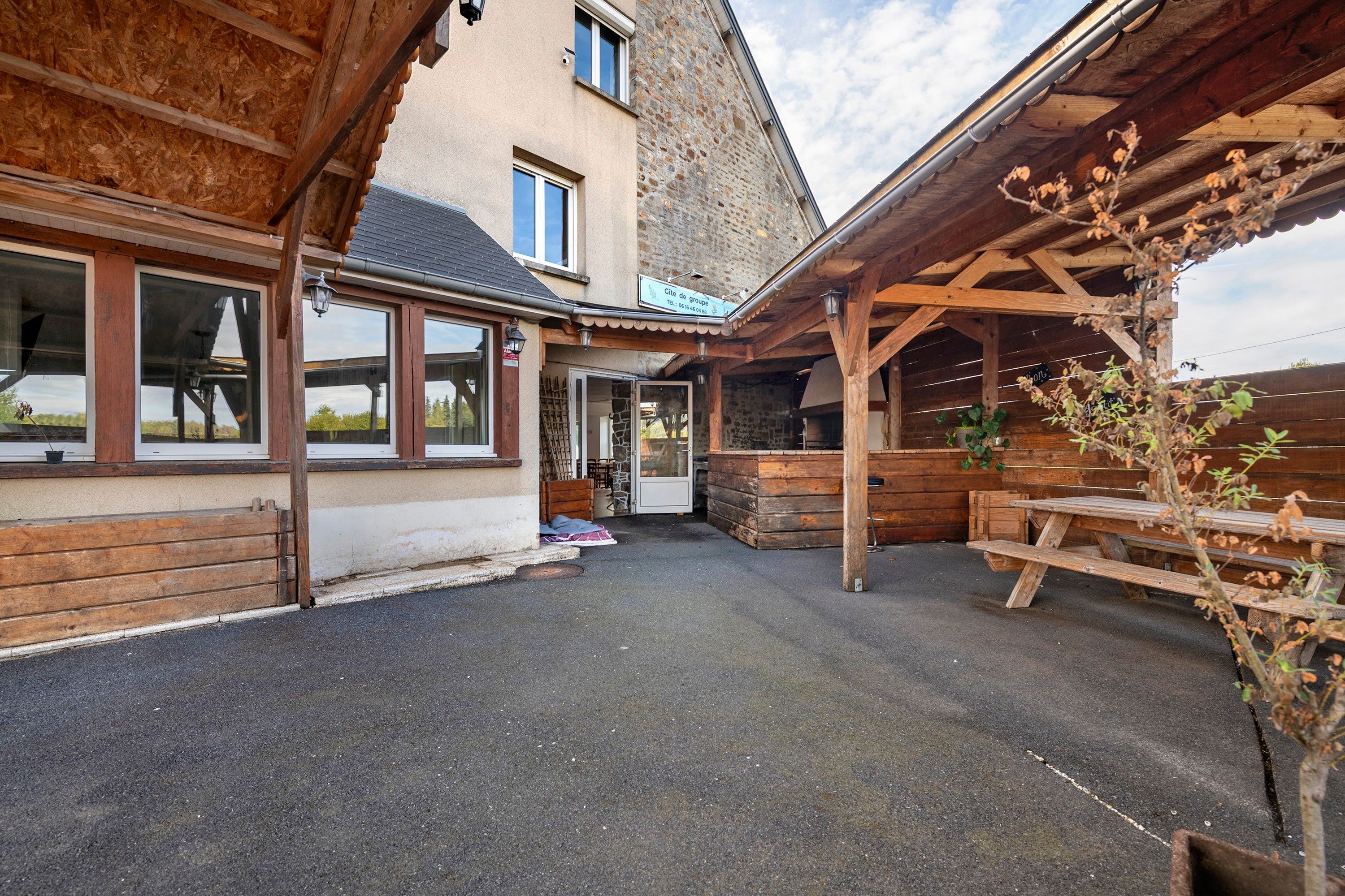 Un espace extérieur abrité, avec un banc en bois et une table, un mur en pierre et un bâtiment doté de grandes fenêtres et d’une porte.