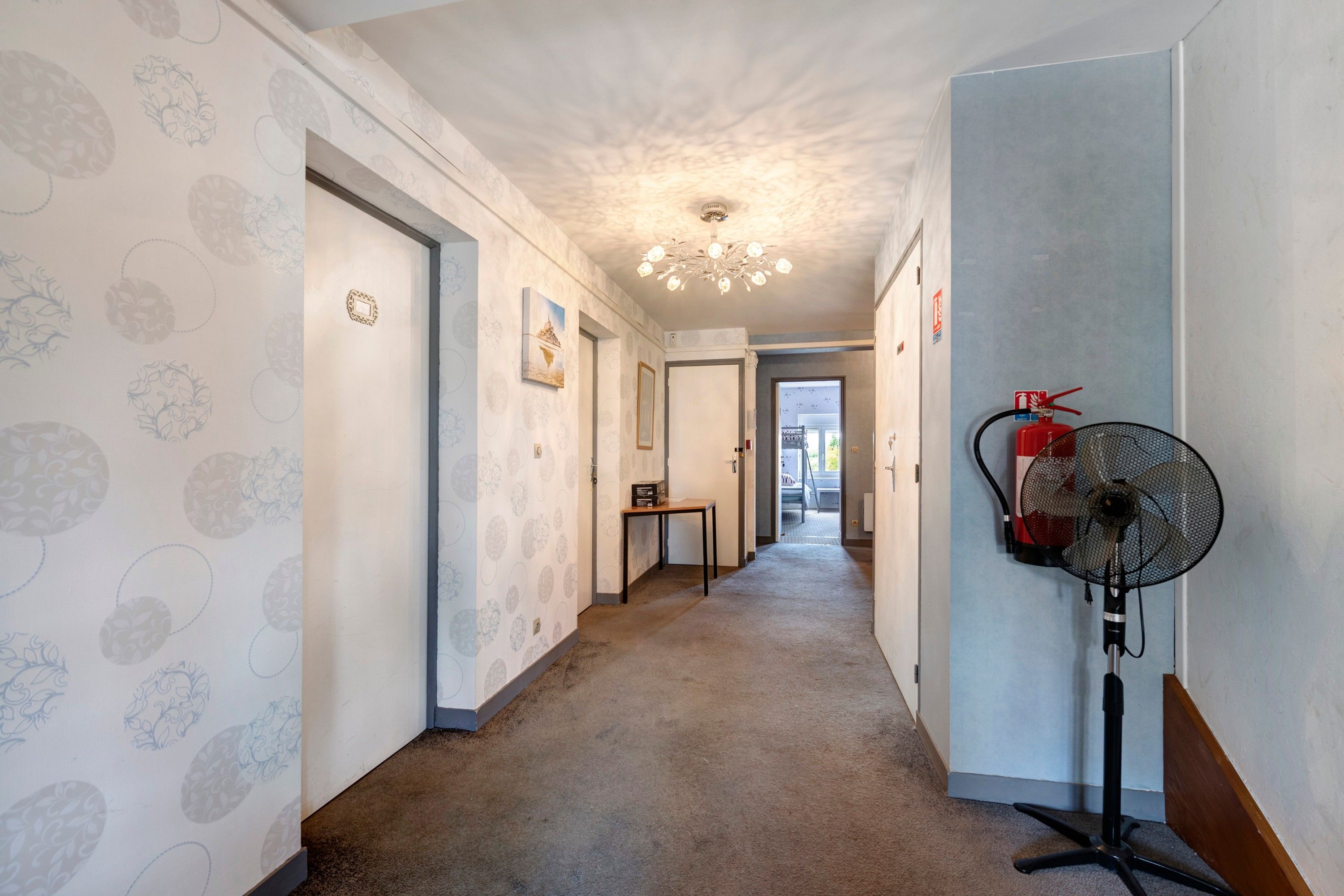 A hotel hallway with patterned wallpaper, a chandelier, a fire extinguisher, and a standing fan.
