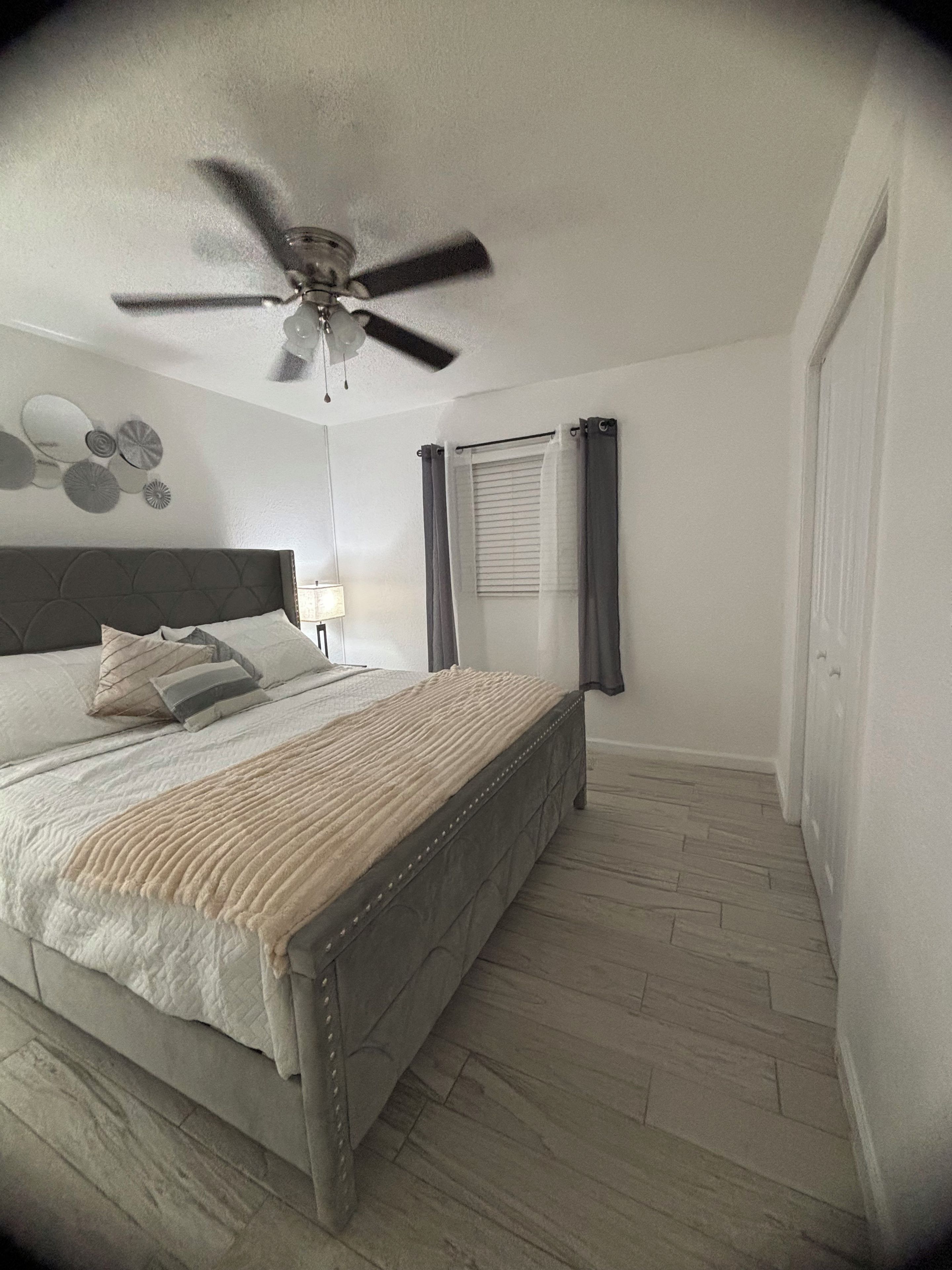 A bedroom with a bed, a ceiling fan, a window with curtains, and a wall-mounted light.